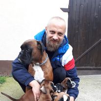 A man with a beard and a colorful jacket in his arms is holding a German boxer and a puppy